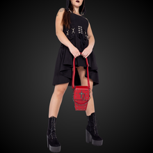 Load image into Gallery viewer, A gothic styled model with dark vampy red makeup wearing a goth black hooded dress holding the GothX Red Mini Coffin Bag between their legs on a black background. The coffin bag is made with vegan friendly sleek red leather and features a detachable decorative metal chain with metal cross studding.