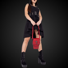 Load image into Gallery viewer, A gothic styled model with dark vampy red makeup wearing a goth black hooded dress holding the GothX Red Mini Coffin Bag between their legs on a black background. The coffin bag is made with vegan friendly sleek red leather and features a detachable decorative metal chain with metal cross studding.
