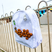 Load image into Gallery viewer, The White &amp; Clear Window Ita Backpack hanging on a metal railing outside a skatepark. The bag is facing forward to highlight the clear front window filled with glittery pumpkins and two metal D rings with a keyring attached, two side elasticated pockets, main zip compartment and top handle. The vegan friendly bag is inspired by kawaii jfashion.
