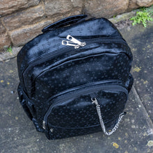 Load image into Gallery viewer, The Black Embossed Cross Backpack sat outside in front of a brick wall in a tumblr grunge style. The backpack is facing forward to highlight the two front silver zip pockets, two elasticated side pockets, main double zip compartment and a silver decorative detachable silver chain draping across the front. The bag is made of vegan friendly leather with 3d embossed crosses in varying sizes all over.