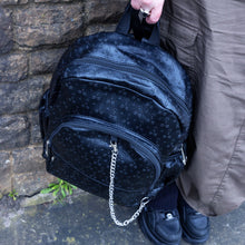 Load image into Gallery viewer, The Black Embossed Cross Backpack being held by a goth grunge model in front of a brick wall in a tumblr grunge style. The backpack is facing forward to highlight the two front silver zip pockets, two elasticated side pockets, main double zip compartment and a silver decorative detachable silver chain draping across the front. The bag is made of vegan friendly leather with 3d embossed crosses in varying sizes all over.