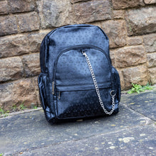 Load image into Gallery viewer, The Black Embossed Cross Backpack sat outside in front of a brick wall in a tumblr grunge style. The backpack is facing forward to highlight the two front silver zip pockets, two elasticated side pockets, main double zip compartment and a silver decorative detachable silver chain draping across the front. The bag is made of vegan friendly leather with 3d embossed crosses in varying sizes all over.