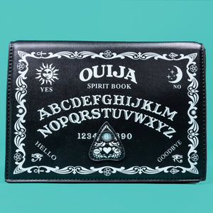 The GothX Ouija Spirit Book Mini Bag sat on a teal background. The bag is forward to highlight the ouija spirit book white printed detailing on the black book bag with the 3D planchette stitching. Bag is inspired by witchy style and necromancy.