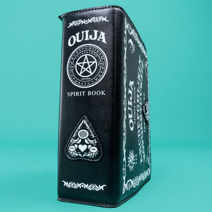 The GothX Ouija Spirit Book Mini Bag sat on a teal background. The bag is forward to highlight the ouija spirit book white printed detailing on the black book bag spine and front with the 3D planchette stitching. Bag is inspired by witchy style and necromancy.