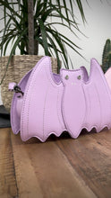 Load and play video in Gallery viewer, The GothX Pastel Lilac Bat Bag and the GothX Pastel Pink Bat Bag sat next to each other on a wooden table with green and black plants behind them. They are both facing forward to highlight the white stitching detailing, 3d bat shape and the silver and white faux crystal eyes.