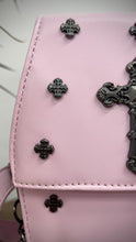 Load and play video in Gallery viewer, The GothX Pastel Pink Mini Coffin Bag sat on a wooden table in front of green and black plants. The bag is facing forward to highlight the cross studs, cross & chain centrepiece, detachable chain and detachable adjustable shoulder strap.