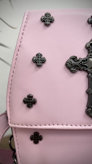 The GothX Pastel Pink Mini Coffin Bag sat on a wooden table in front of green and black plants. The bag is facing forward to highlight the cross studs, cross & chain centrepiece, detachable chain and detachable adjustable shoulder strap.