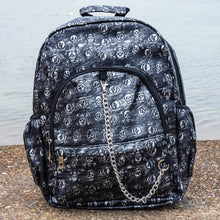 Load image into Gallery viewer, The Rustic Skulls Backpack sat on a grunge gritty concrete floor in front of misty water. The bag is facing forward to highlight the two front zip pockets, two elasticated side pockets, main zip compartment, the top handle and the detachable decorative silver chain. All over the backpack is an embossed 3d texture skulls and skull and crossbones on a faux leather material in a brushed black and silver grunge style.
