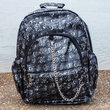 Load image into Gallery viewer, The Rustic Skulls Backpack sat on a grunge gritty concrete floor in front of misty water. The bag is facing forward to highlight the two front zip pockets, two elasticated side pockets, main zip compartment, the top handle and the detachable decorative silver chain. All over the backpack is an embossed 3d texture skulls and skull and crossbones on a faux leather material in a brushed black and silver grunge style.