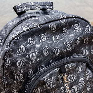 The Rustic Skulls Backpack sat on a concrete floor. A close up of the bag top handle and double zip opening main compartment. All over the backpack is an embossed 3d texture skulls and skull and crossbones on a faux leather material in a brushed black and silver grunge style.