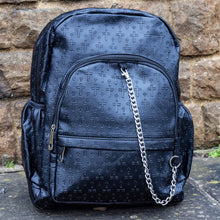 Load image into Gallery viewer, The Black Embossed Cross Backpack sat outside in front of a brick wall in a tumblr grunge style. The backpack is facing forward to highlight the two front silver zip pockets, two elasticated side pockets, main double zip compartment and a silver decorative detachable silver chain draping across the front. The bag is made of vegan friendly leather with 3d embossed crosses in varying sizes all over.