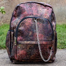Load image into Gallery viewer, The Rustic Skulls &amp; Roses Backpack sat outside in front of a warm toned grunge wall and grass. The bag is facing forward to highlight the two zip front pockets, two elasticated side pockets, the double zip main compartment with a silver draping chain across the front. The vegan friendly faux leather bag is varying colours of bronze, gold, rose and brown with black 3d embossed sugar skulls, roses and swirls all over.
