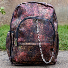 Load image into Gallery viewer, The Rustic Skulls & Roses Backpack sat outside in front of a warm toned grunge wall and grass. The bag is facing forward to highlight the two zip front pockets, two elasticated side pockets, the double zip main compartment with a silver draping chain across the front. The vegan friendly faux leather bag is varying colours of bronze, gold, rose and brown with black 3d embossed sugar skulls, roses and swirls all over.
