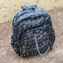 Load image into Gallery viewer, The Rustic Skulls Backpack sat on a grunge gritty concrete floor in front of misty water. The bag is facing forward to highlight the two front zip pockets, two elasticated side pockets, main zip compartment, the top handle and the detachable decorative silver chain. All over the backpack is an embossed 3d texture skulls and skull and crossbones on a faux leather material in a brushed black and silver grunge style.