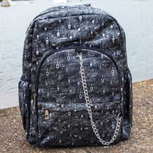 Load image into Gallery viewer, The Rustic Silver Spider Backpack sat outside on a gritty concrete floor with misty water behind. The bag is facing forward to highlight the two zip front pockets, two elasticated side pockets, the double zip main compartment with a silver draping chain across the front. The vegan friendly faux leather bag has 3d embossed spiders in varying sizes with brushed black and silver tones all over.
