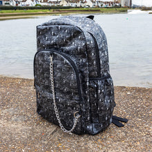 Load image into Gallery viewer, The Rustic Silver Spider Backpack sat outside on a gritty concrete floor with misty water behind. The bag is facing forward angled left to highlight the two zip front pockets, two elasticated side pockets, the double zip main compartment with a silver draping chain across the front. The vegan friendly faux leather bag has 3d embossed spiders in varying sizes with brushed black and silver tones all over.