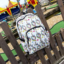 Load image into Gallery viewer, The Rainbow Unicorn Backpack hanging off a wooden fence in front of a multicoloured fairground area by the beach. The vegan friendly backpack is a white canvas material with repeating kawaii cute unicorn heads with pink blue and yellow manes with hearts surrounding them. The bag is facing forward to highlight the front two zip pockets, the main zip compartment and silver draping decorative chain.