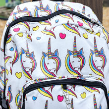 Load image into Gallery viewer, Close up of the Rainbow Unicorn Backpack hanging off a wooden fence in front of a multicoloured fairground area by the beach. The vegan friendly backpack is a white canvas material with repeating kawaii cute unicorn heads with pink blue and yellow manes with hearts surrounding them. The bag is facing forward to highlight the front two zip pockets, the main zip compartment and silver draping decorative chain.