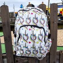 Load image into Gallery viewer, The Rainbow Unicorn Backpack hanging off a wooden fence in front of a multicoloured fairground area by the beach. The vegan friendly backpack is a white canvas material with repeating kawaii cute unicorn heads with pink blue and yellow manes with hearts surrounding them. The bag is facing forward to highlight the front two zip pockets, the main zip compartment and silver draping decorative chain.