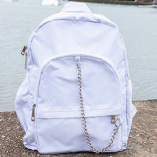 Load image into Gallery viewer, The white backpack with chain is sat outside on concrete floor at a waters edge. The vegan friendly bag is facing forward to highlight the top handle, the double zip main compartment, two front zip pockets with a silver decorative detachable draping chain and two elasticated side pockets.