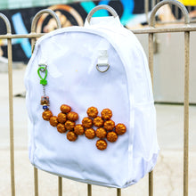 Load image into Gallery viewer, The White &amp; Clear Window Ita Backpack hanging on a metal railing outside a skatepark. The bag is facing forward to highlight the clear front window filled with glittery pumpkins and two metal D rings with a keyring attached, two side elasticated pockets, main zip compartment and top handle. The vegan friendly bag is inspired by kawaii jfashion.

