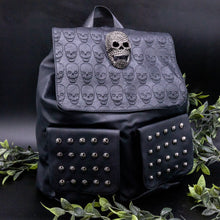 Load image into Gallery viewer, GothX twin pocket skull vegan backpack on a black background with leaves surrounding it. The bag is facing forward to highlight the diamante effect skull, skull embossed vegan leather front flap, tassel tie cords and two silver studded front pockets.