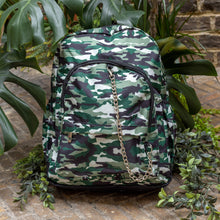 Load image into Gallery viewer, Classic Camouflage vegan backpack with chain sat outside in front of a tropical plant and brick wall. The backpack with a green and brown camo print is facing forward highlighting the two front zip pockets, two side pockets and silver decorative chain.