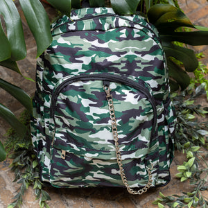 Classic Camouflage vegan backpack with chain sat outside in front of a tropical plant and brick wall. The backpack with a green and brown camo print is facing forward highlighting the two front zip pockets, two side pockets and silver decorative chain.