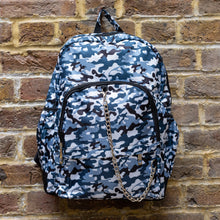 Load image into Gallery viewer, The Snow Camouflage Backpack hanging outside on a brick wall. The white, blue, navy and black vegan friendly backpack is facing forward to highlight the two front zip pockets with a silver draping detachable chain, the two side elasticated pockets, the top handle and the main double zip compartment.
