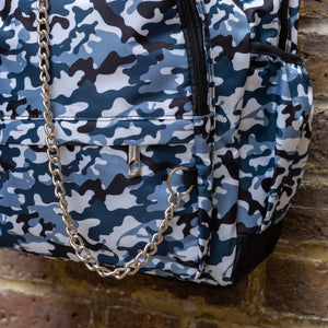 Close up of The Snow Camouflage Backpack hanging outside on a brick wall. The white, blue, navy and black vegan friendly backpack is facing forward to highlight the two front zip pockets with a silver draping detachable chain, the two side elasticated pockets, the top handle and the main double zip compartment.