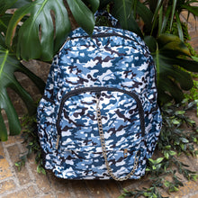 Load image into Gallery viewer, The Snow Camouflage Backpack sat outside on a brick floor surrounded by giant monstera leaves and other green foliage. The white, blue, navy and black vegan friendly camo backpack is sat facing forward to highlight the two front zip pockets with a silver draping detachable chain, the two side elasticated pockets, the top handle and the main double zip compartment.