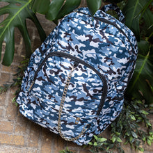 Load image into Gallery viewer, The Snow Camouflage Backpack sat outside on a brick floor surrounded by giant monstera leaves and other green foliage. The white, blue, navy and black vegan friendly camo backpack is sat facing forward to highlight the two front zip pockets with a silver draping detachable chain, the two side elasticated pockets, the top handle and the main double zip compartment.