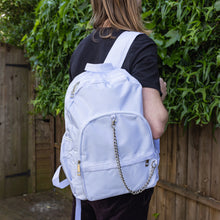 Load image into Gallery viewer, The white backpack with chain is being held by a blonde male model's shoulder. The vegan friendly bag is facing forward to highlight the top handle, the double zip main compartment, two front zip pockets with a silver decorative detachable draping chain and two elasticated side pockets.