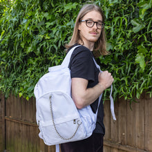 Load image into Gallery viewer, The white backpack with chain is being held by a blonde male model's shoulder whilst they look over their shoulder to camera. The vegan friendly bag is facing forward to highlight the top handle, the double zip main compartment, two front zip pockets with a silver decorative detachable draping chain and two elasticated side pockets.