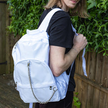 Load image into Gallery viewer, The white backpack with chain is being held by a blonde male model's shoulder whilst they look over their shoulder to right floor. The vegan friendly bag is facing forward to highlight the top handle, the double zip main compartment, two front zip pockets with a silver decorative detachable draping chain and two elasticated side pockets.