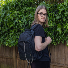 Load image into Gallery viewer, Jack is stood outside in a garden area wearing the black nylon vegan backpack with chain on his back whilst looking over his shoulder to camera. The photo is cropped from the thighs up. The backpack is all black with two front zip pockets, two side pockets and a silver decorative chain across the front to the side.