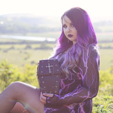 Load image into Gallery viewer, Alternative purple haired goth model sat on a grass field in a goth outfit holding the GothX Mini Coffin Vegan Cross Body Bag to camera. The vegan leather bag is facing forward angled to the right to highlight the detachable chain, cross studded front and stud cross centrepiece with chain.