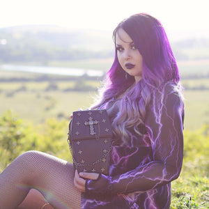 Alternative purple haired goth model sat on a grass field in a goth outfit holding the GothX Mini Coffin Vegan Cross Body Bag to camera. The vegan leather bag is facing forward angled to the right to highlight the detachable chain, cross studded front and stud cross centrepiece with chain.