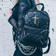 Load image into Gallery viewer, The GothX Quilted Cross Vegan Mini Backpack on the shoulder of a model wearing a black mesh top and black skirt. The mini bag has adjustable straps, quilted front and gunmetal coloured metal detailing with a cross appliqué. 
