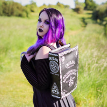 Load image into Gallery viewer, The gothx ouija spirit book vegan backpack modelled by Azariah. They are facing away from the camera with the backpack on their back facing the camera to highlight the embroidered planchette and white printed detailing featuring a ouija board, pentagrams and lace pattern.
