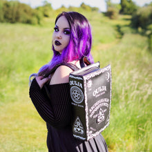 Load image into Gallery viewer, The gothx ouija spirit book vegan backpack modelled by Azariah. They are facing away from the camera with the backpack on their back facing the camera to highlight the embroidered planchette and white printed detailing featuring a ouija board, pentagrams and lace pattern.