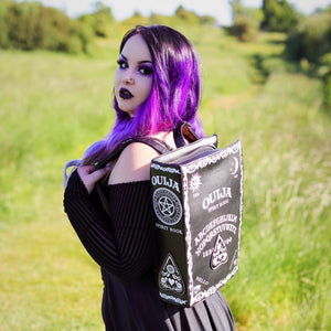 The gothx ouija spirit book vegan backpack modelled by Azariah. They are facing away from the camera with the backpack on their back facing the camera to highlight the embroidered planchette and white printed detailing featuring a ouija board, pentagrams and lace pattern.