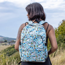 Load image into Gallery viewer, Meera is stood in a grass field wearing the kawaii unicorn vegan backpack on her back. The backpack is facing the camera, highlighting the pastel blue rainbow unicorn print, two front zip pockets, two side pockets and detachable silver chain.
