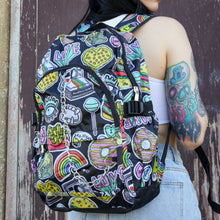 Load image into Gallery viewer, The kawaii graffiti doodle vegan backpack being worn by a tattooed model in front of a brown painted wall. The bag is facing forward to highlight the two front zip pockets, two elastic side pockets & silver chain. The all over print has lollipops, pugs, rainbows, pizzas, polaroid cameras, eyes and fries.