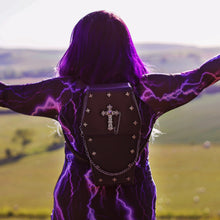 Load image into Gallery viewer, Azariah with long purple gradient hair stood in a field wearing the gothx coffin vegan backpack with a purple lightning dress. They are facing away from the camera with their arms out to their side whilst wearing the backpack on their back. The backpack is facing the camera to highlight the cross mini studs, cross and chain emblem and detachable silver chain.