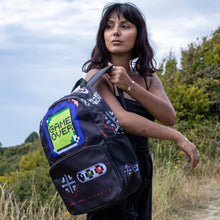 Load image into Gallery viewer, Meera is stood in a grass field wearing an all black outfit with the black game over vegan backpack being put onto her shoulder. The backpack is a black base with a green game over screen with gaming button controls on the front zip pocket, there's two side elasticated net pockets and two zips on the main opening.