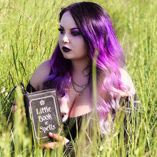 Load image into Gallery viewer, Alternative goth model with black to purple gradient hair sat in a grass field wearing a goth outfit holding the gothx little book of spells vegan shoulder bag open like a book. The witchy inspired vegan leather bag is facing towards the camera to highlight the white printed design of a pentagram, framing and text reading little book of spells.
