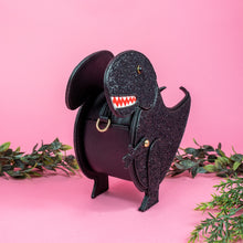 Load image into Gallery viewer, The kawaii black glitter dino vegan bag on a pink studio background with green foliage surrounding it. The bag is angled left and forward to highlight the dinosaur face, moveable arm, detachable strap and black glitter front.