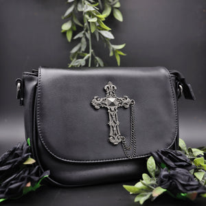 The gothx don't cross me vegan shoulder bag on a black studio background with green foliage and black roses surrounding it. The bag is facing forward to highlight the studded cross chain centrepiece.