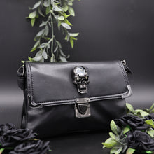 Load image into Gallery viewer, The GothX crystal skull vegan shoulder bag on a black studio background with black roses and green foliage surrounding it. The bag is facing forward to highlight the metal clasp close, metal trim detailing and crystal skull centrepiece.